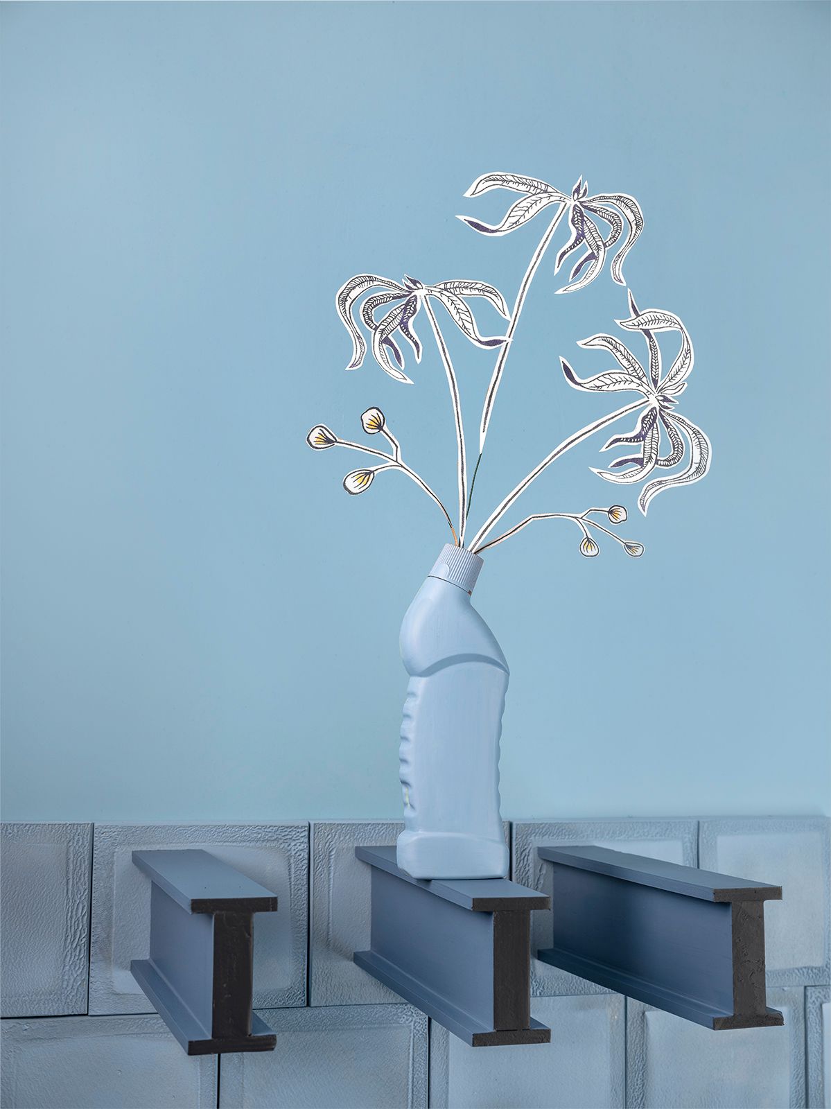 Een decoratief zilverkleurig bloemstuk in een lichtblauwe, hergebruikte fles, geplaatst op metalen balken en betonnen blokken tegen een blauwe achtergrond.