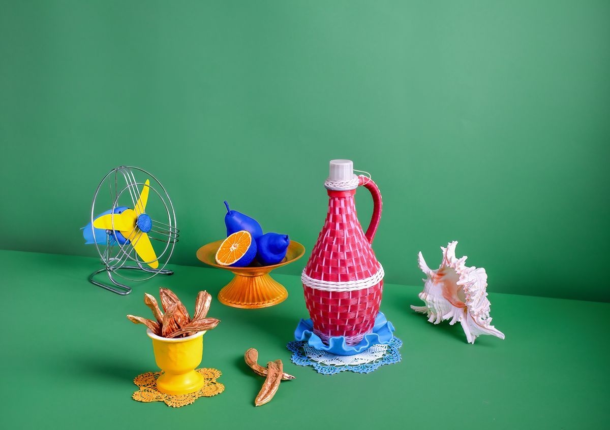 Stilleven met een rode fles, blauw fruit, waaier, schelp en gele versieringen op een groene achtergrond.