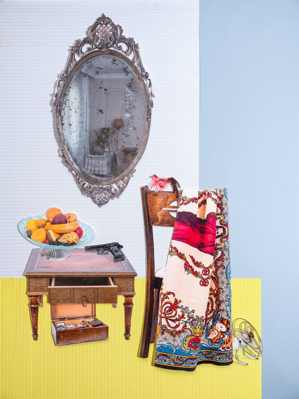 Antieke spiegel, tafel met fruit en andere voorwerpen; deken op een stoel, tegen een gele en blauwe muur.