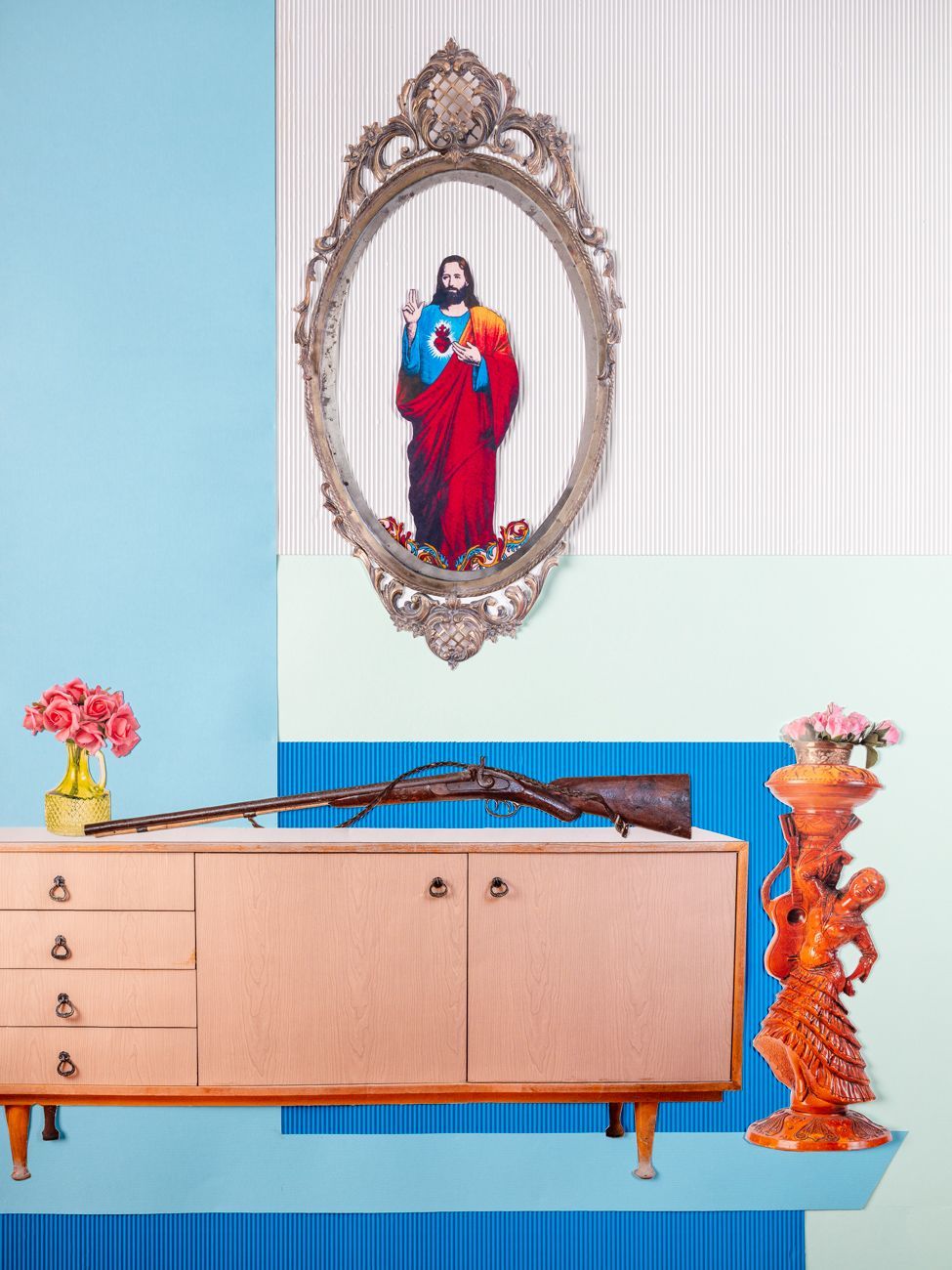Een houten kast met een geweer en een bloemstuk. Een ingelijste afbeelding van Jezus. Blauwe en turquoise achtergrond.