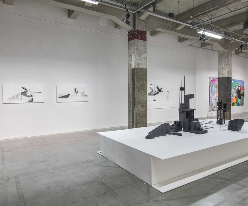 Interieur van een kunstgalerie. Witte muren, betonnen vloer. Sculpturen op een platform, abstracte tekeningen.
