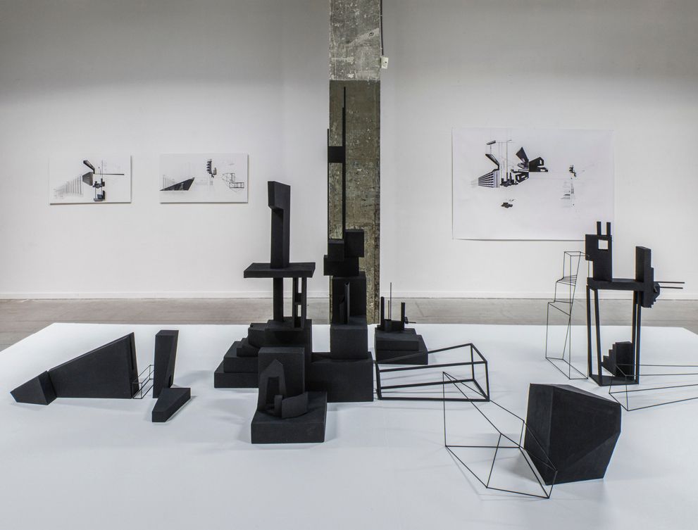 Zwarte geometrische sculpturen en schetsen tentoongesteld in een galerie met witte muren.