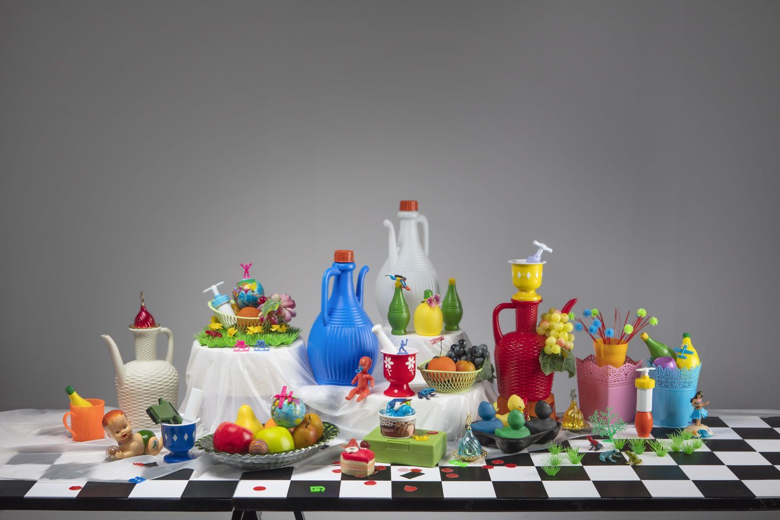 Stilleven met kleurrijke voorwerpen: kannen, taarten en fruit op een geblokte tafel.