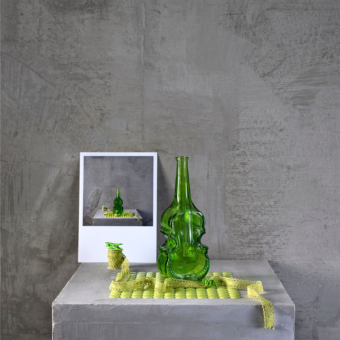 Een kleine sculptuur, een groene glazen fles in de vorm van een viool, ingelijst en geplaatst op een betonnen blok tegen een grijze muur.