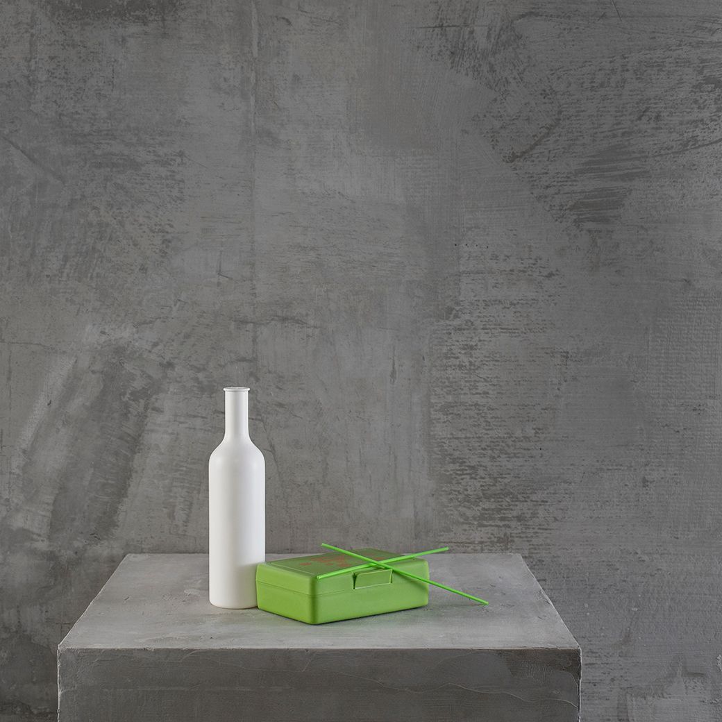 Een witte fles en een groene doos op een grijze vierkante tafel tegen een grijze betonnen muur.