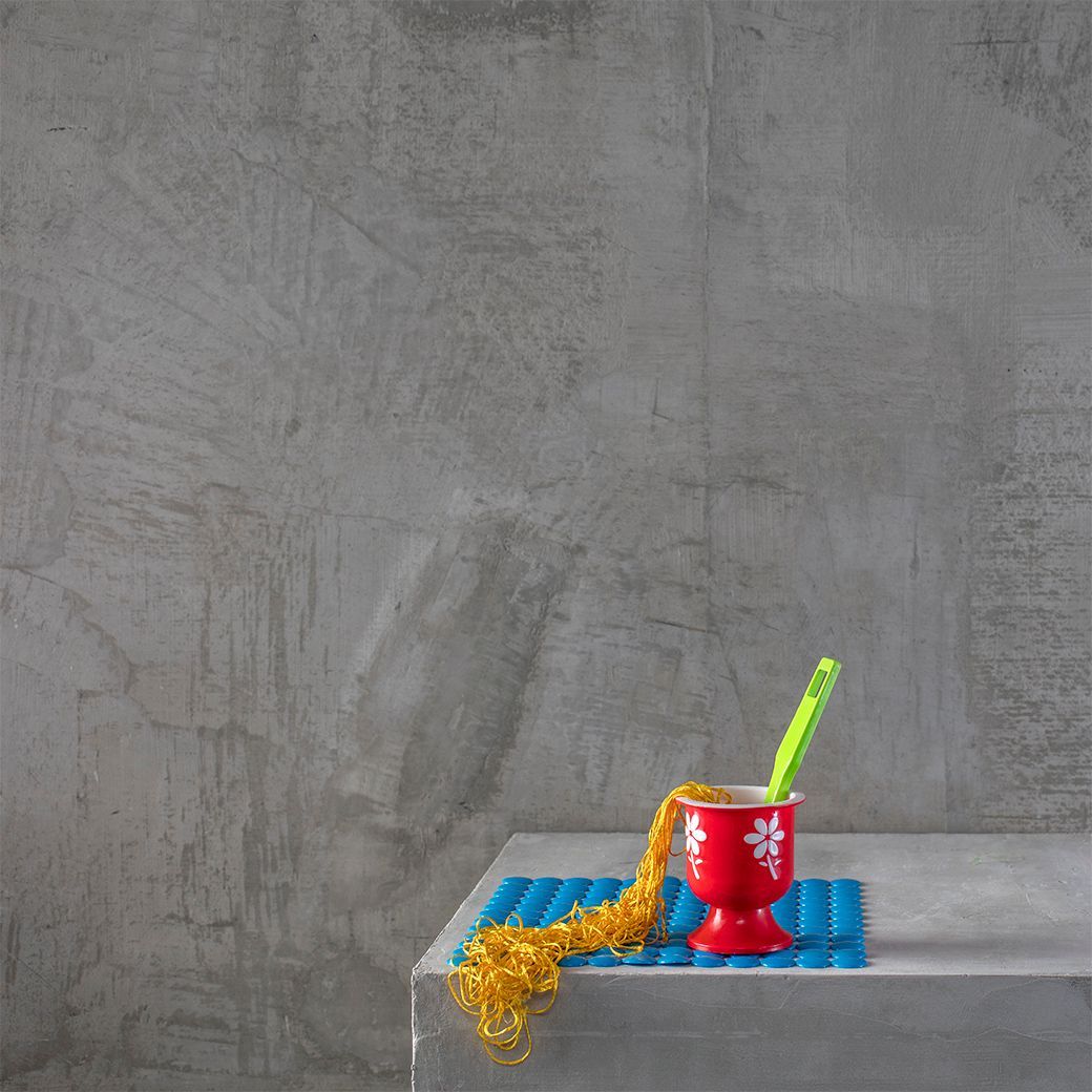 Rode beker met gele noedels en groene lepel op een blauw matje, tegen een grijze muur.
