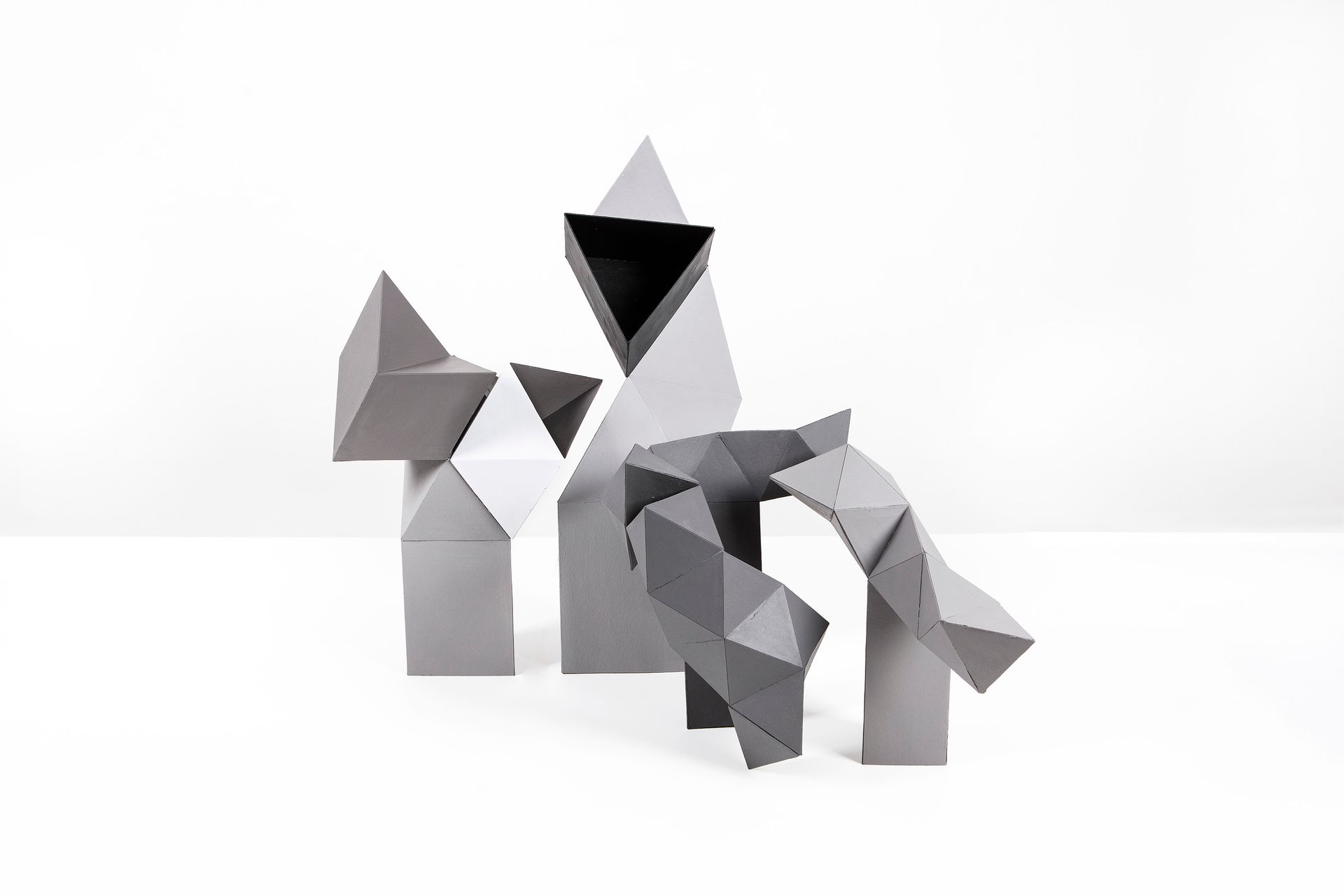 Geometrische, grijze papieren sculpturen op een witte achtergrond, in diverse vormen en maten, die op abstracte figuren lijken.