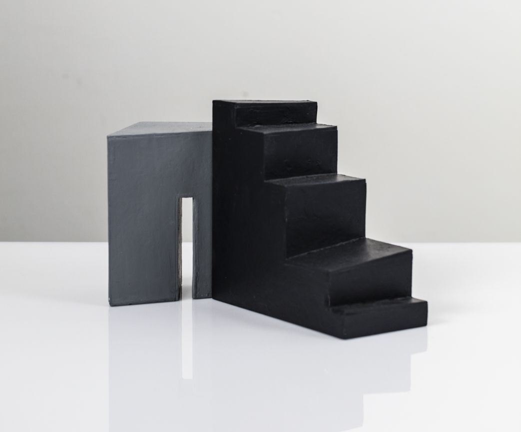 Abstracte sculptuur, grijs blok verbonden met zwarte trapachtige treden, witte achtergrond.