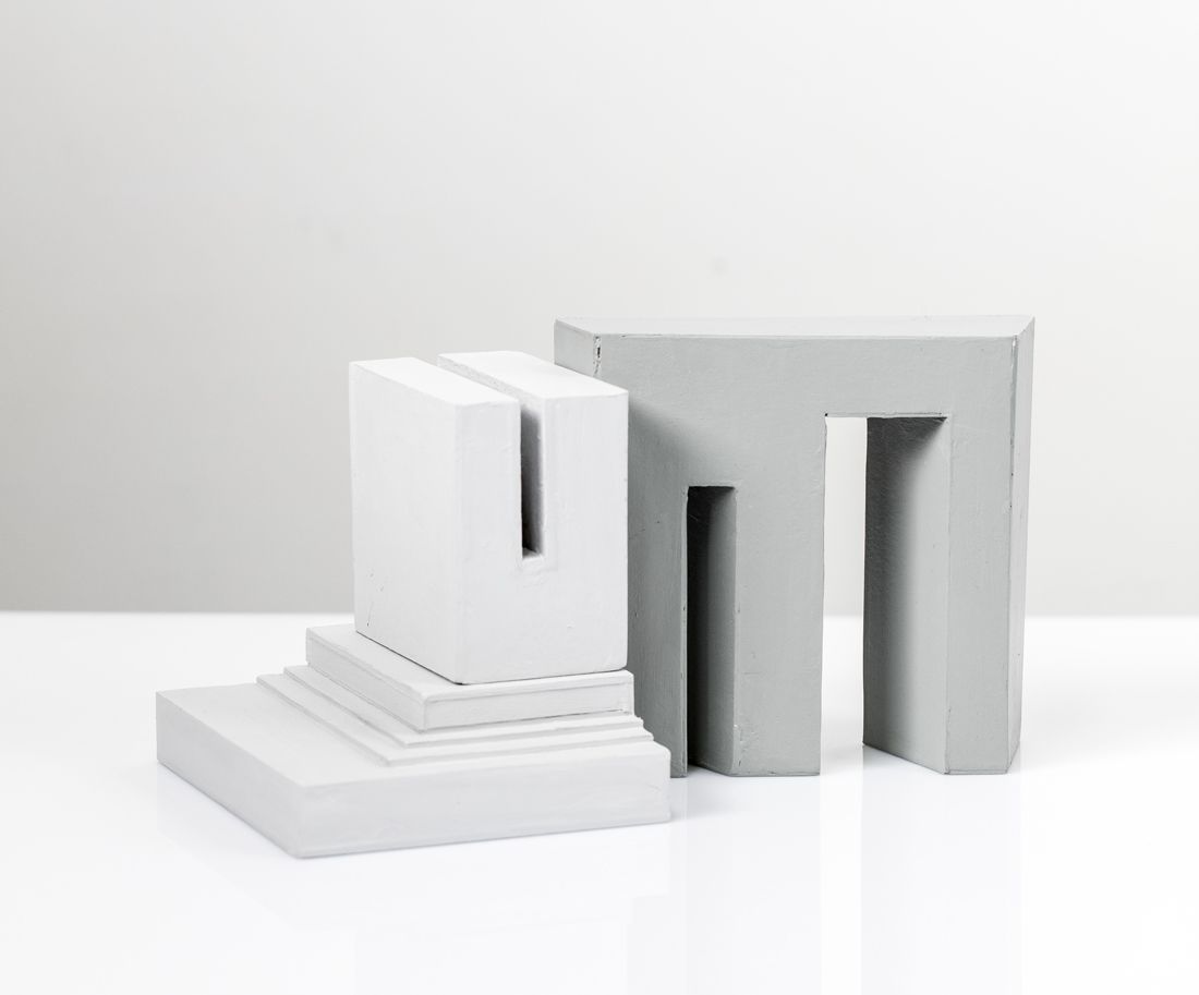 Twee abstracte, geometrische sculpturen, de ene U-vormig, de andere een boogvormige deuropening, in grijstinten, geplaatst op een witte ondergrond.