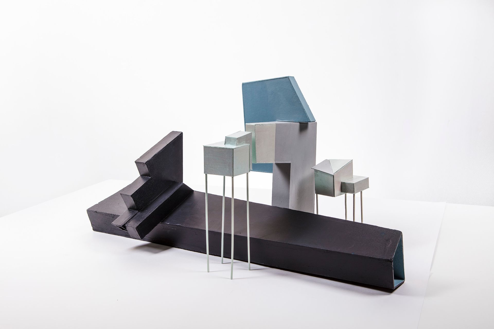Abstracte geometrische sculptuur, zwarte en pastelkleurige blokken op dunne zilveren poten, tegen een witte achtergrond.