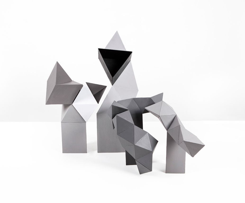 Abstracte, grijze, geometrische papieren sculpturen op een witte ondergrond, met hoekige vormen en schaduwen.