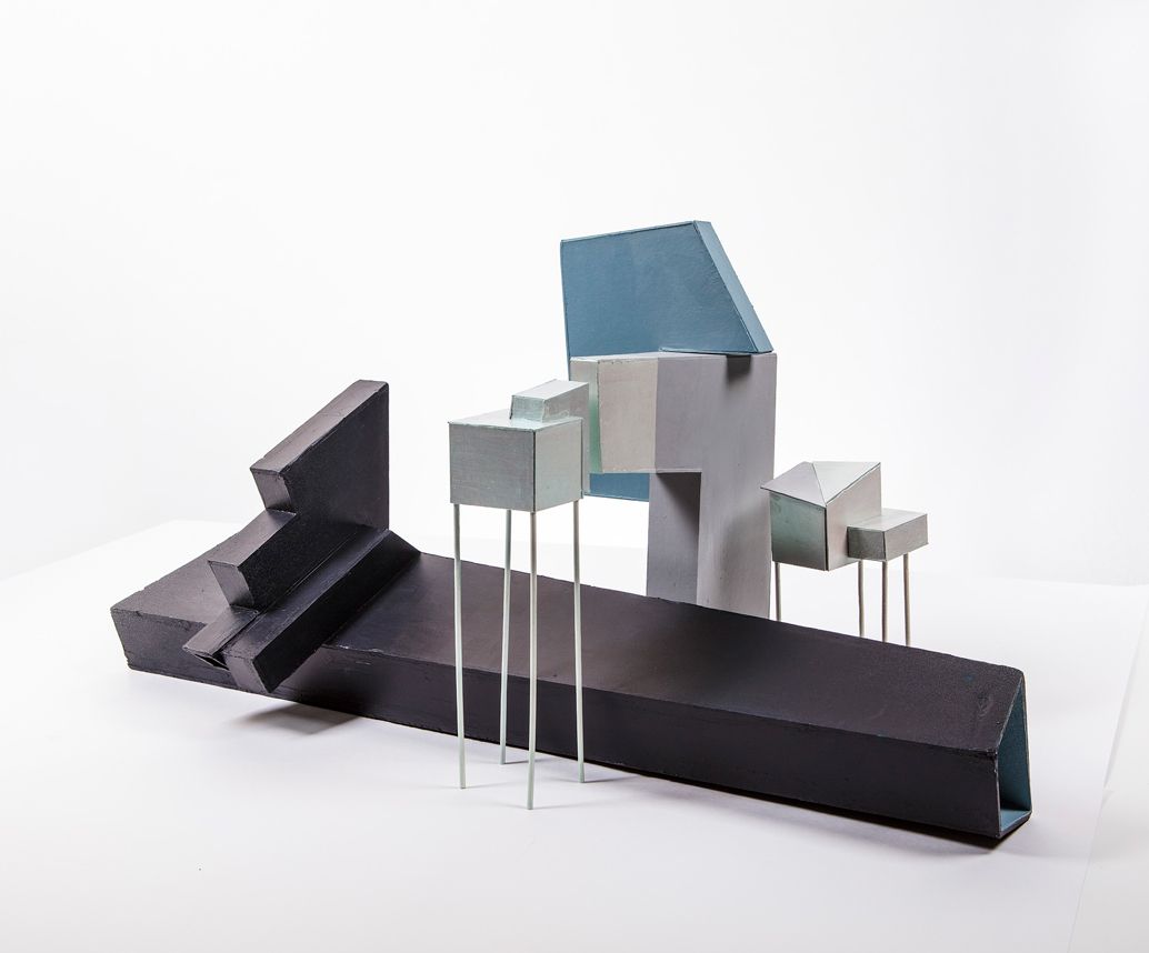 Abstracte sculptuur van geometrische vormen, voornamelijk zwart, grijs en blauw, sommige op dunne metalen poten, tegen een witte achtergrond.