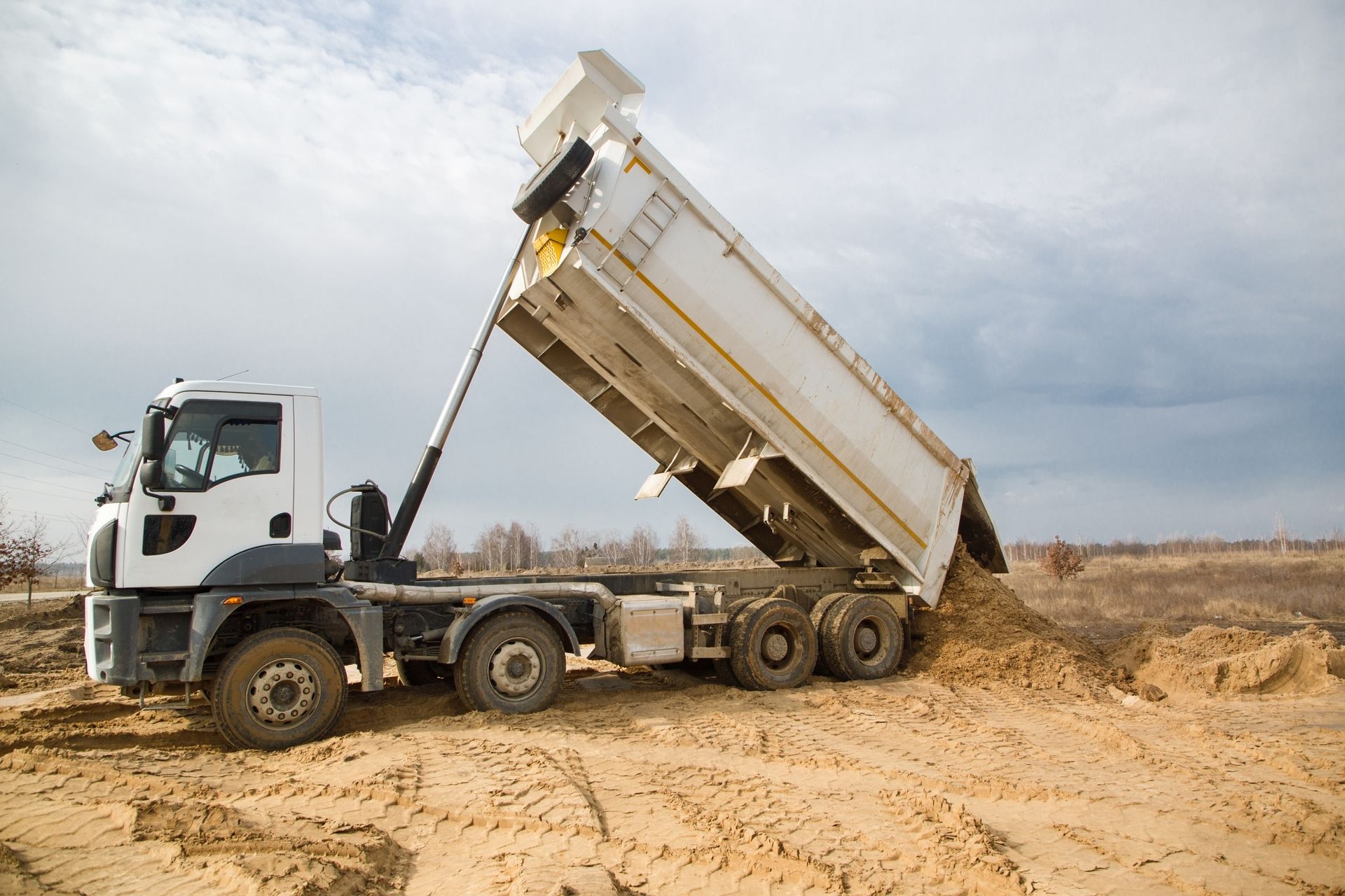 White dump truck unloading dirt on a sandy ground under a cloudy sky. White dump truck unloading dirt on a sandy ground under a cloudy sky.
