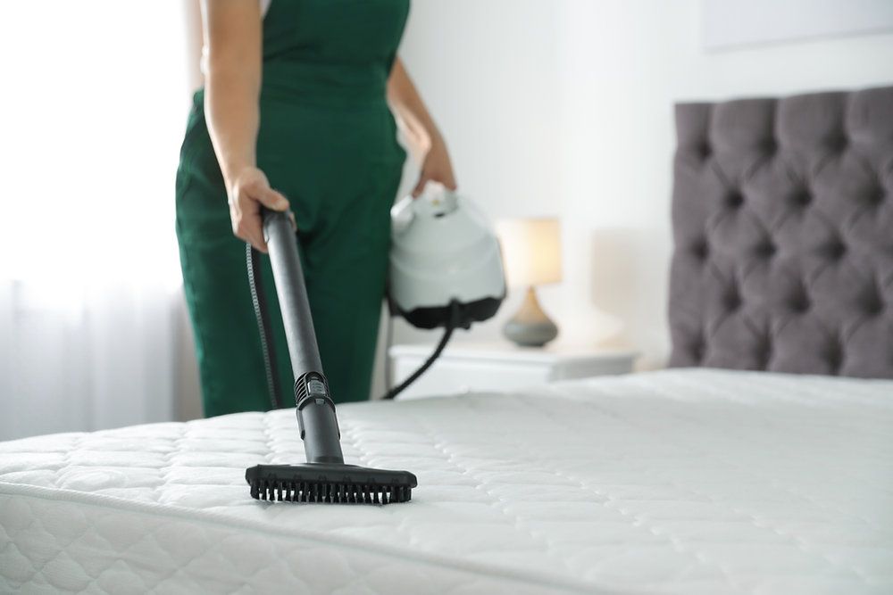 Person vacuuming a white mattress in a bright bedroom with a green outfit