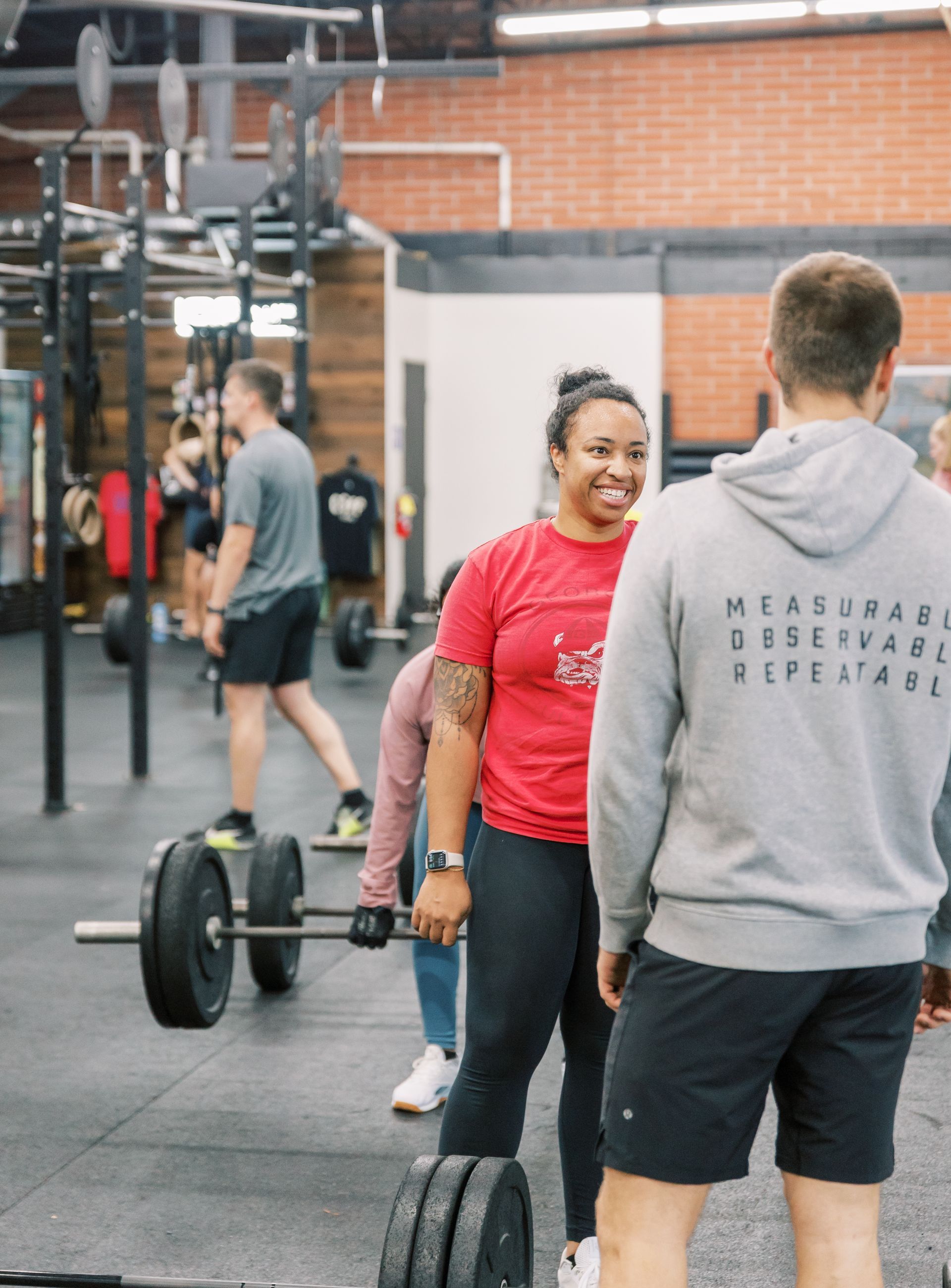 Two people smiling in a gym, with barbells and weight plates on the floor.