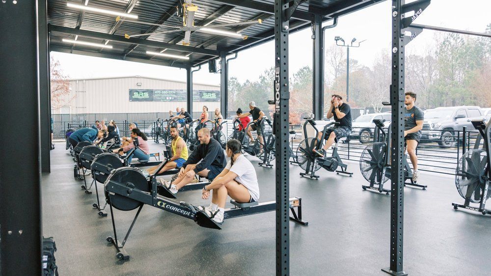 People exercising in a gym with rowing machines and other cardio equipment under an open metal roof.
