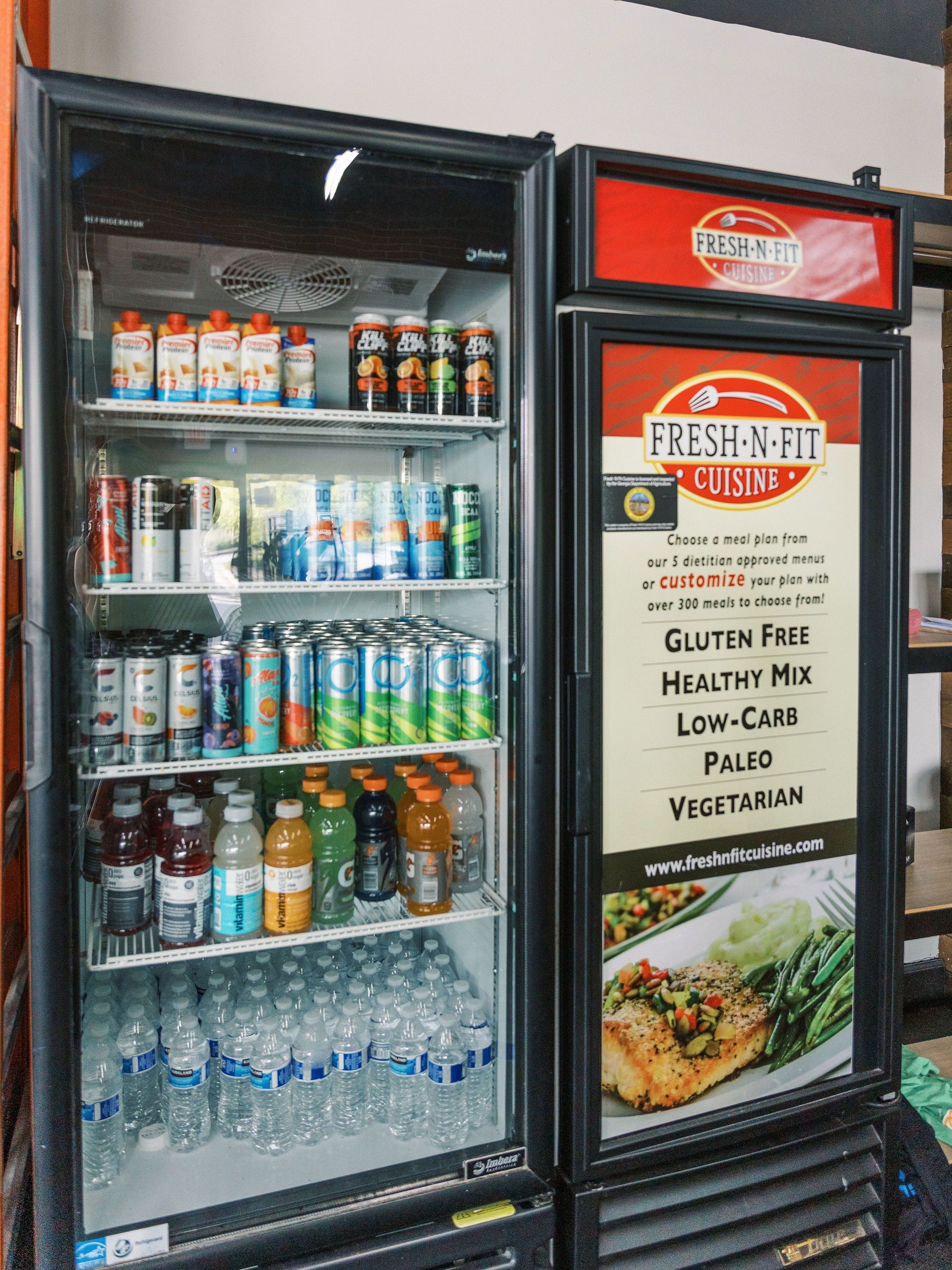 Open glass beverage cooler beside a Fresh Up restaurant menu board with drinks and sandwiches displayed.