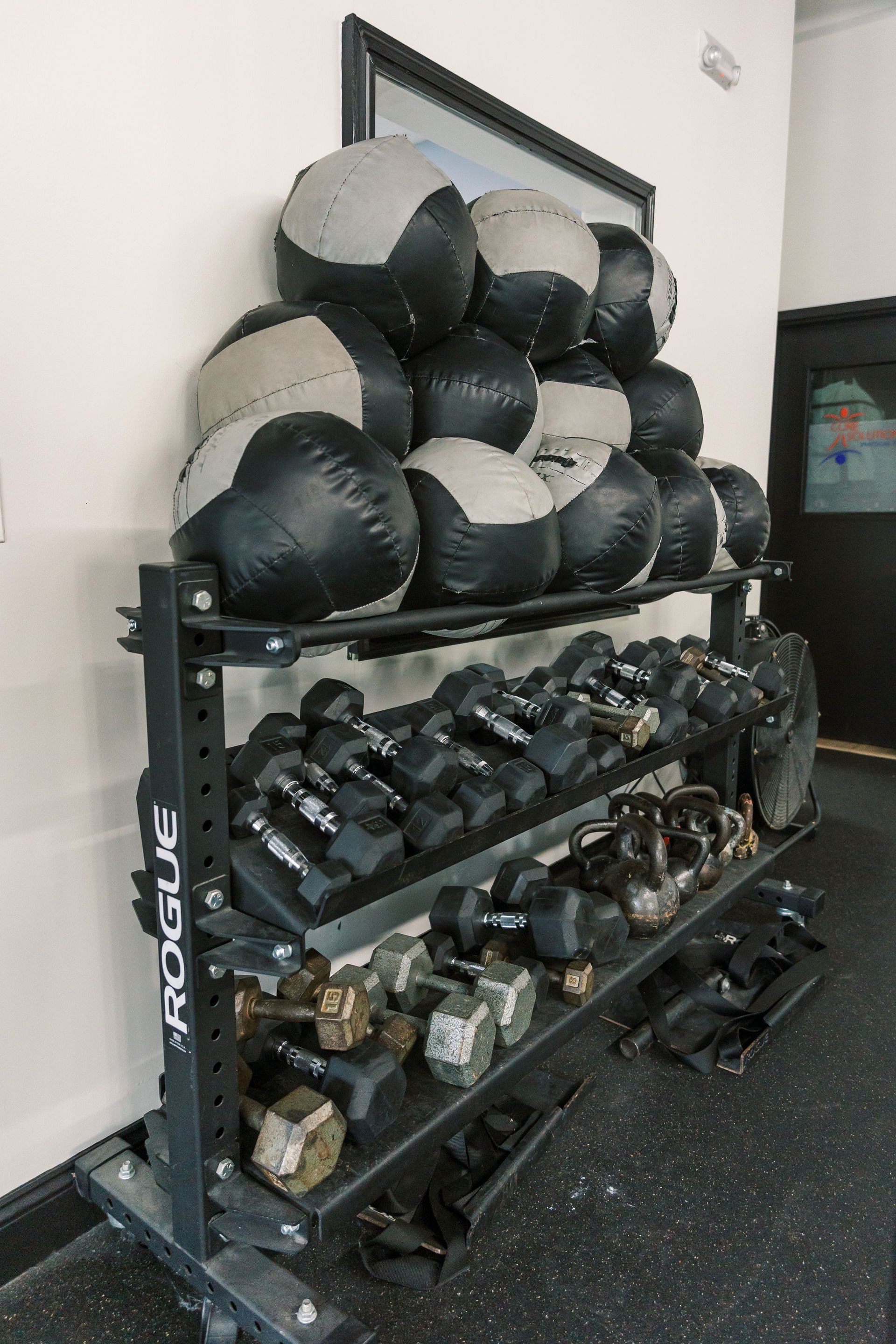Gym rack with medicine balls, dumbbells, and kettlebells against a white wall