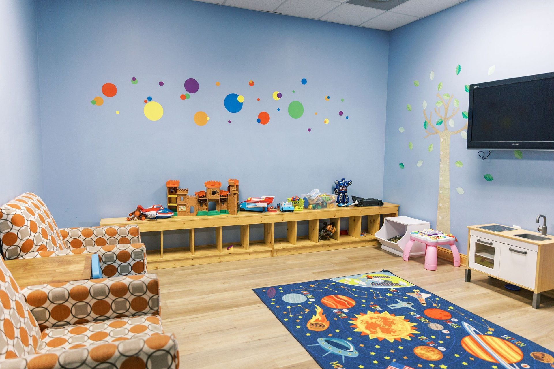 Playroom with colorful wall decals, toys on low shelves, patterned chairs, rug, and wall-mounted TV.