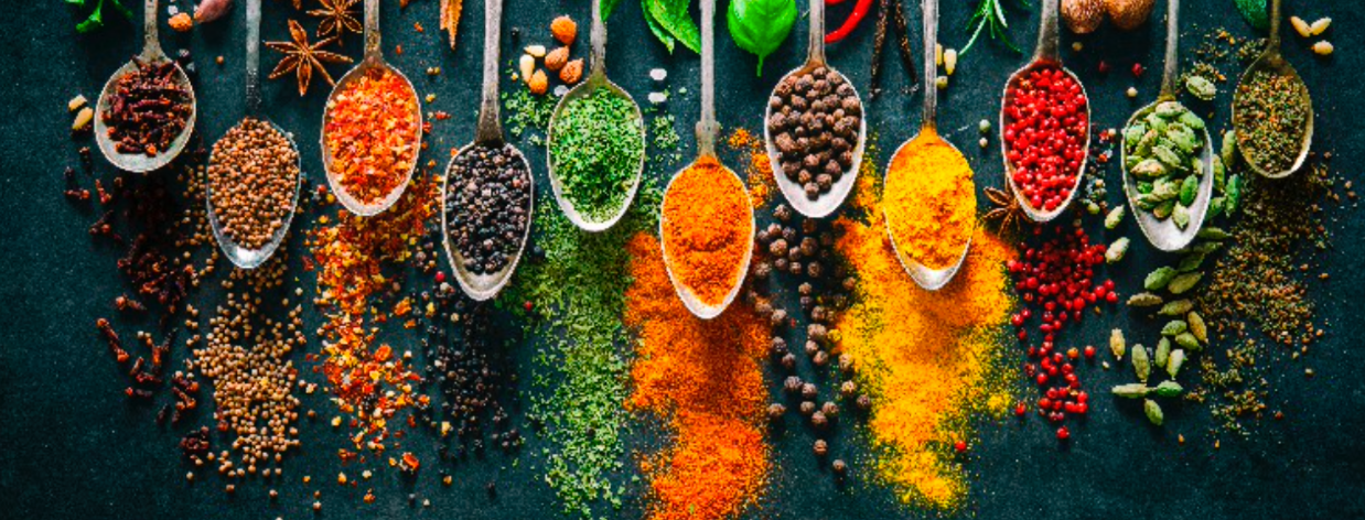Assortment of colorful spices in silver spoons arranged on a dark surface.