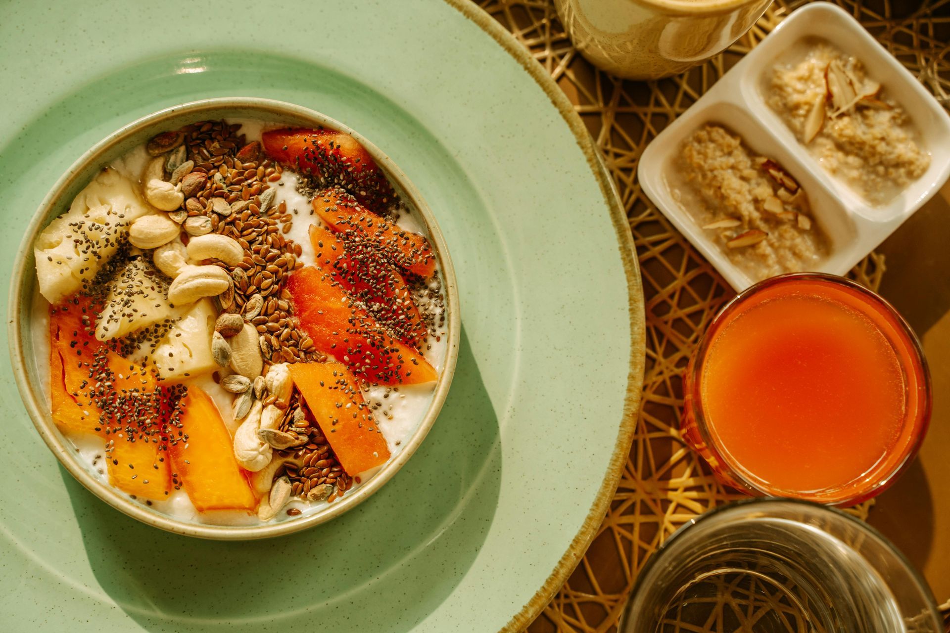 Breakfast bowl with fruit, nuts, and seeds on a green plate, beside a juice and side dish.