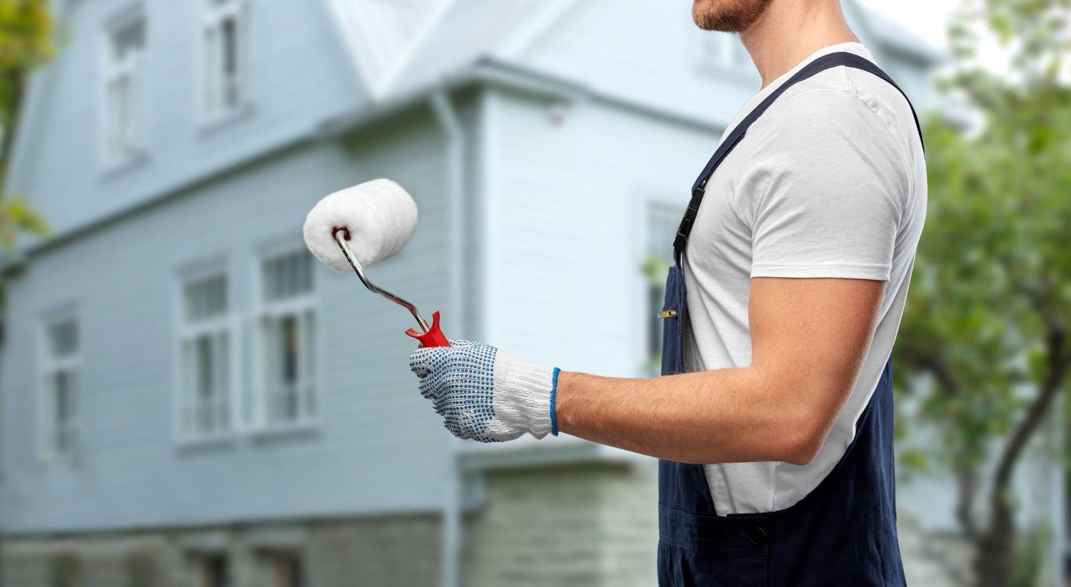 Worker in gloves applying white paint to a house exterior with a roller