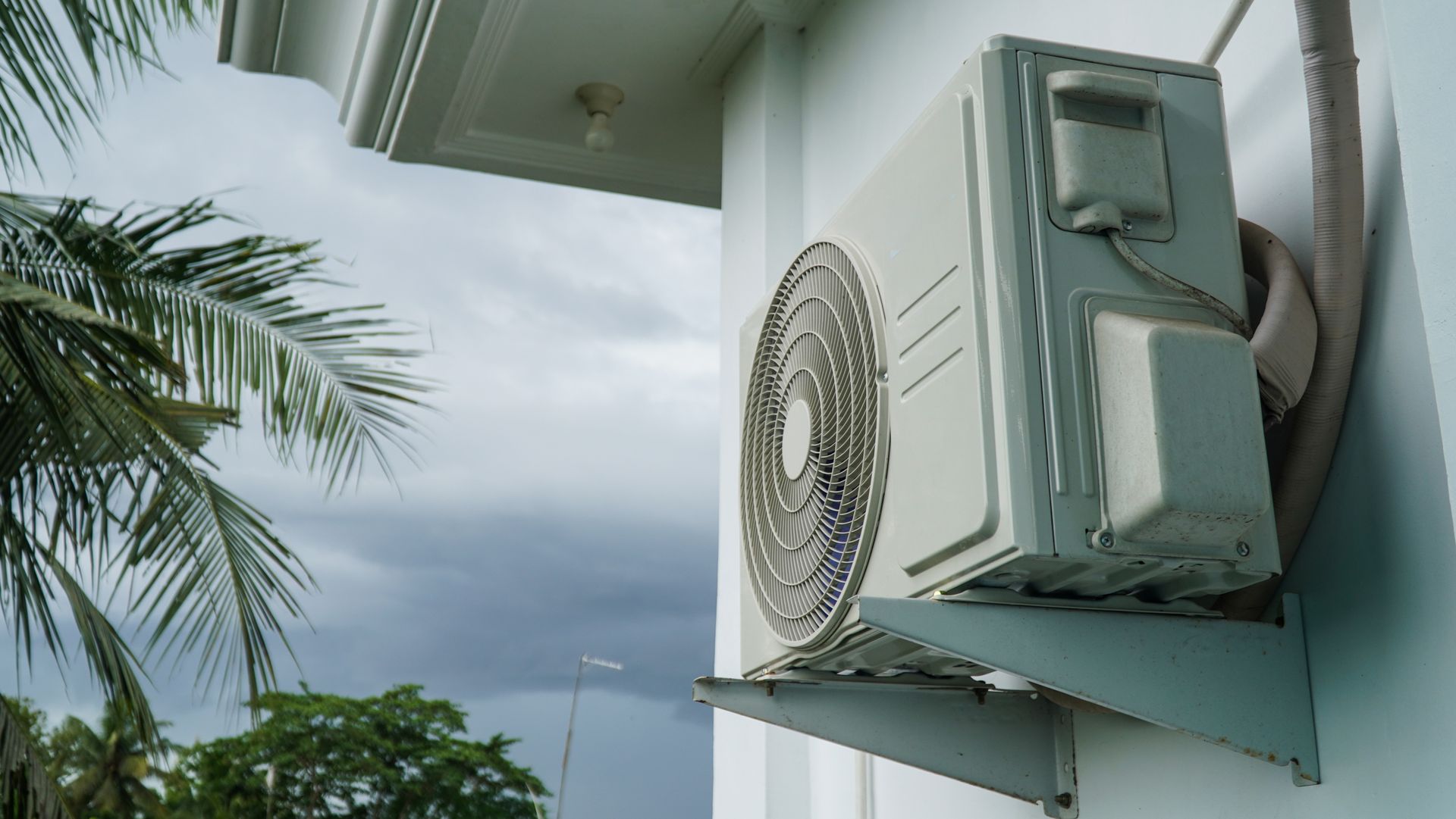An outdoor air conditioning unit mounted to a white wall under the edge of a roof with palm trees visible in the background.