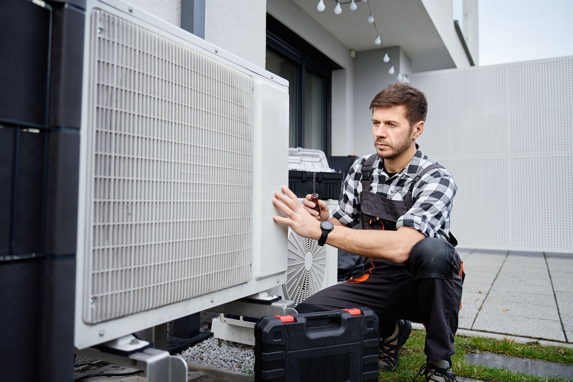 A technician in workwear kneeling to inspect and repair an outdoor air conditioning unit with a tool case nearby.