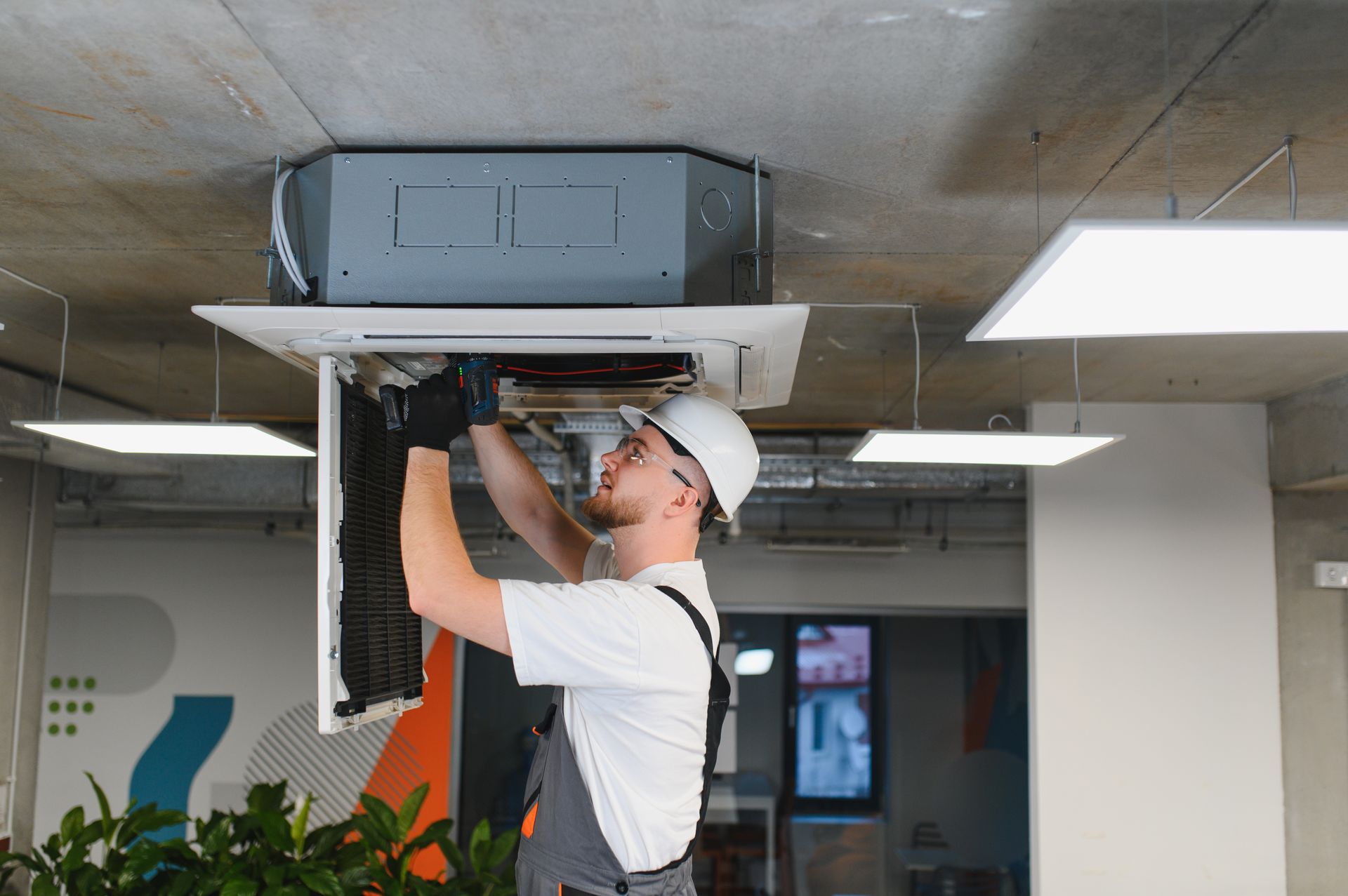 A technician in a hard hat repairs a ceiling-mounted air conditioning unit in an office.