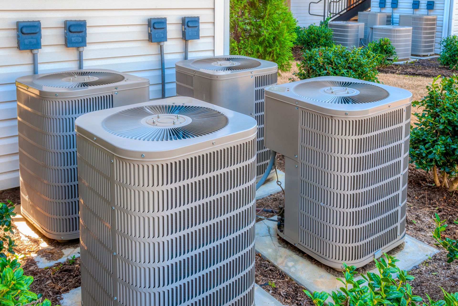 Three gray air conditioning units sit on concrete pads beside a building wall, surrounded by mulch and green bushes.