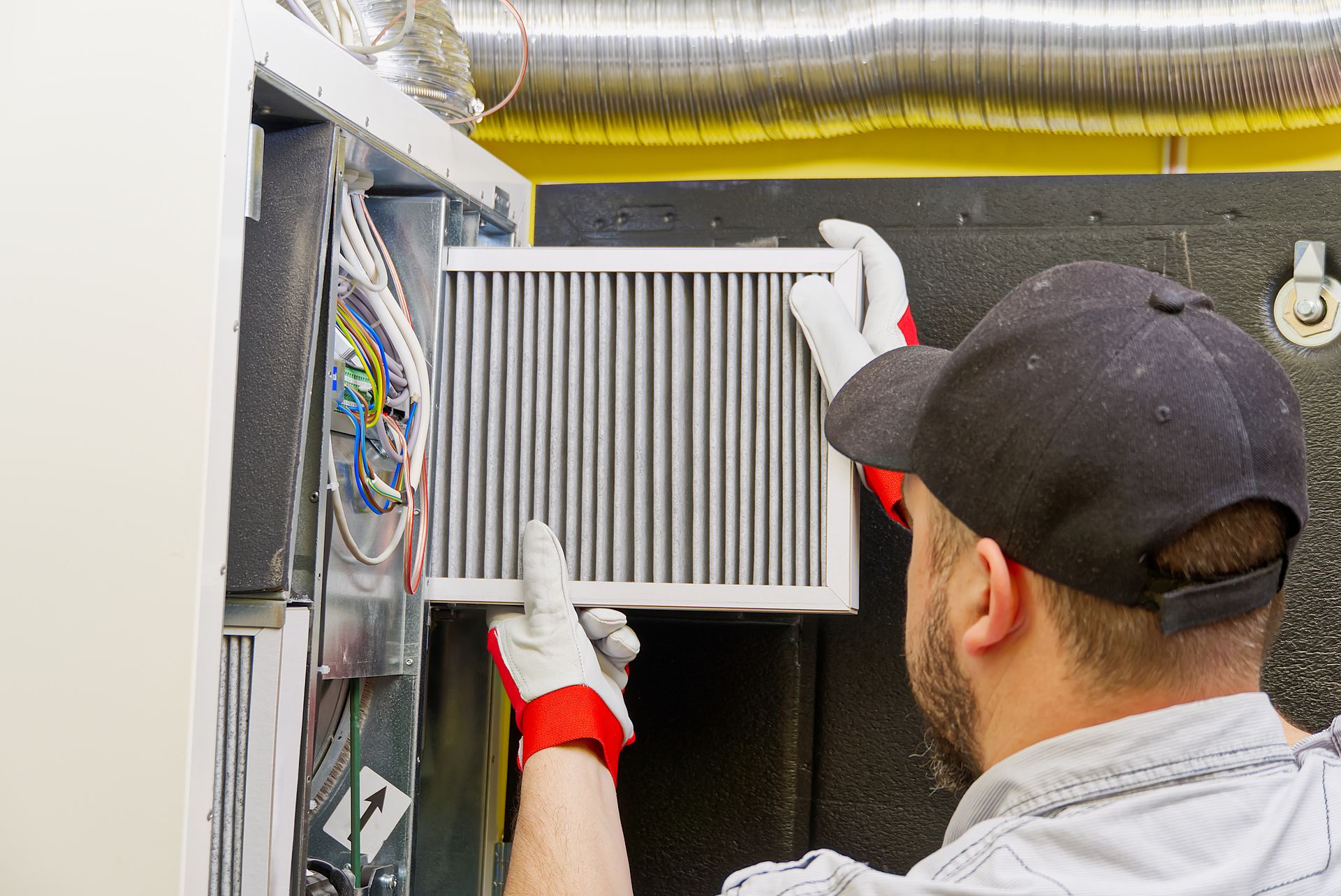A technician in gloves and a baseball cap installs a rectangular HVAC filter into a furnace unit.