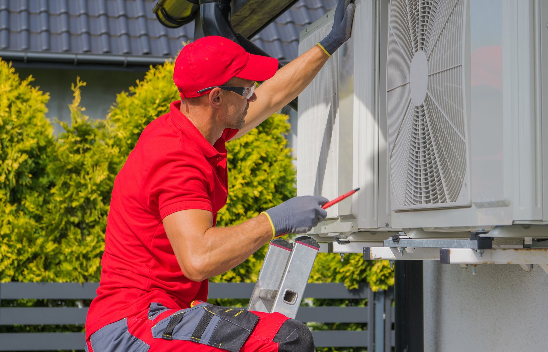 A technician in a red uniform and protective eyewear uses a screwdriver to service an outdoor HVAC unit near a green hedge.