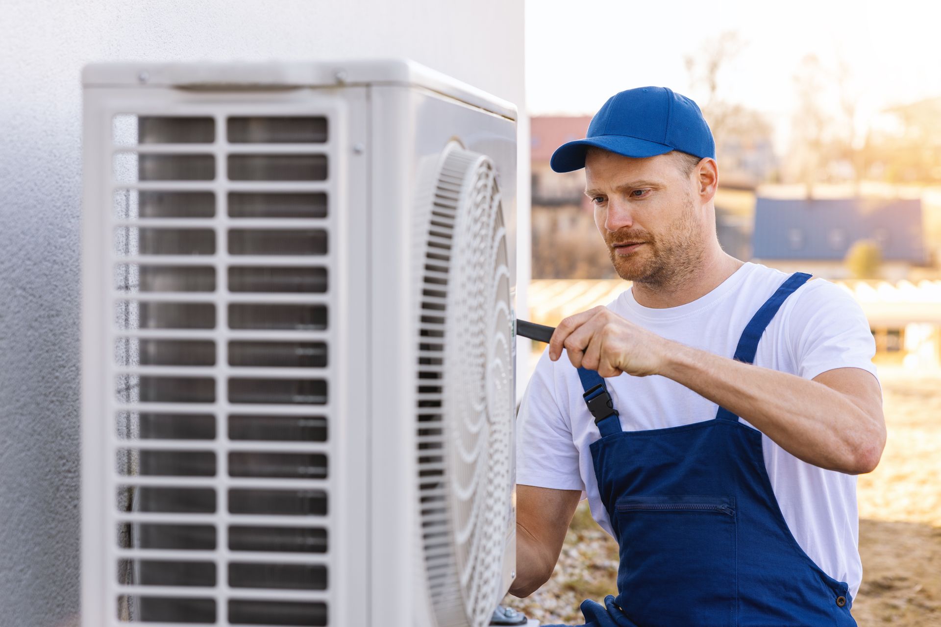 A technician wearing a blue cap and work overalls repairs an outdoor air conditioning unit with a screwdriver.