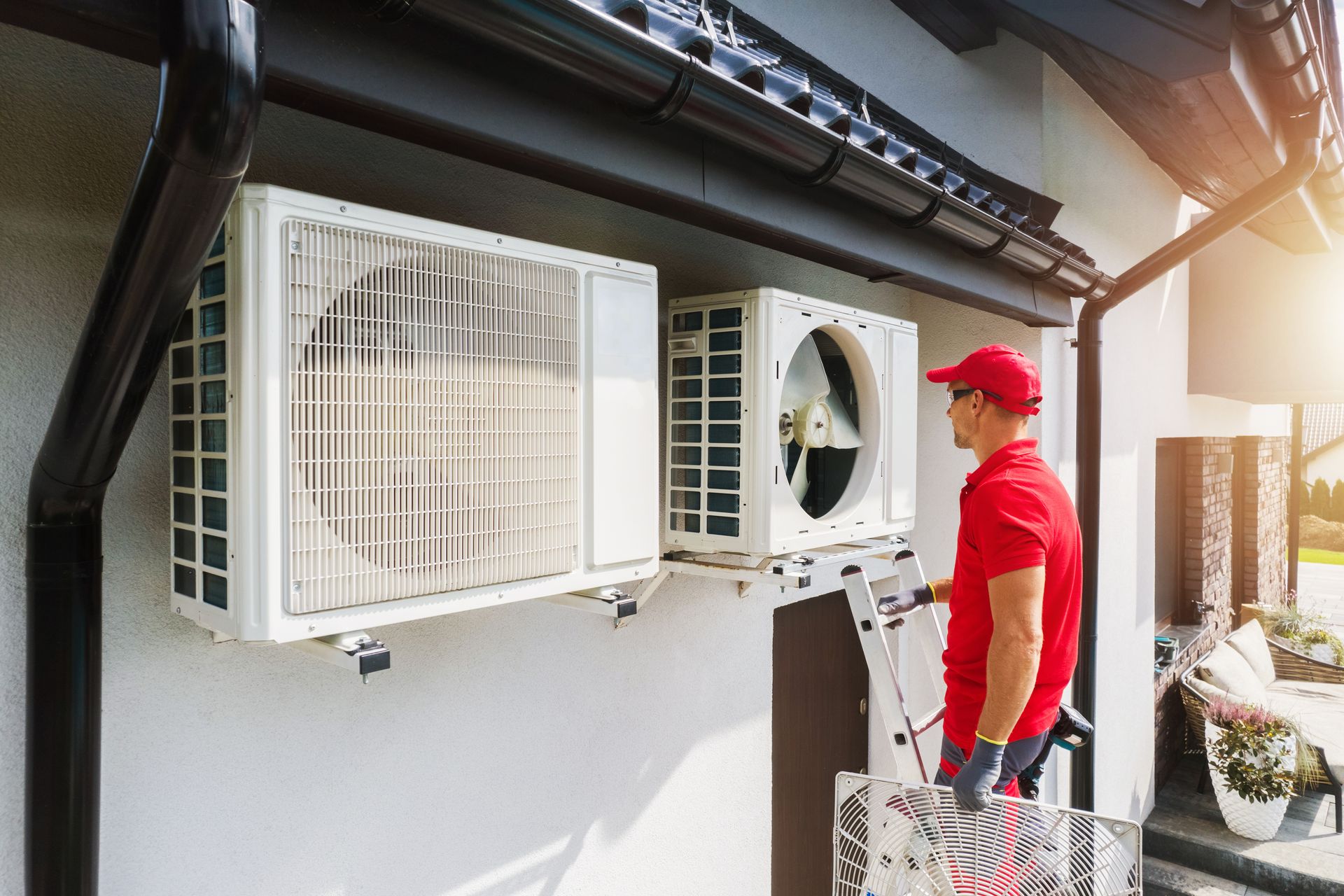 A technician in a red uniform installs or repairs an air conditioning unit on the exterior wall of a house.