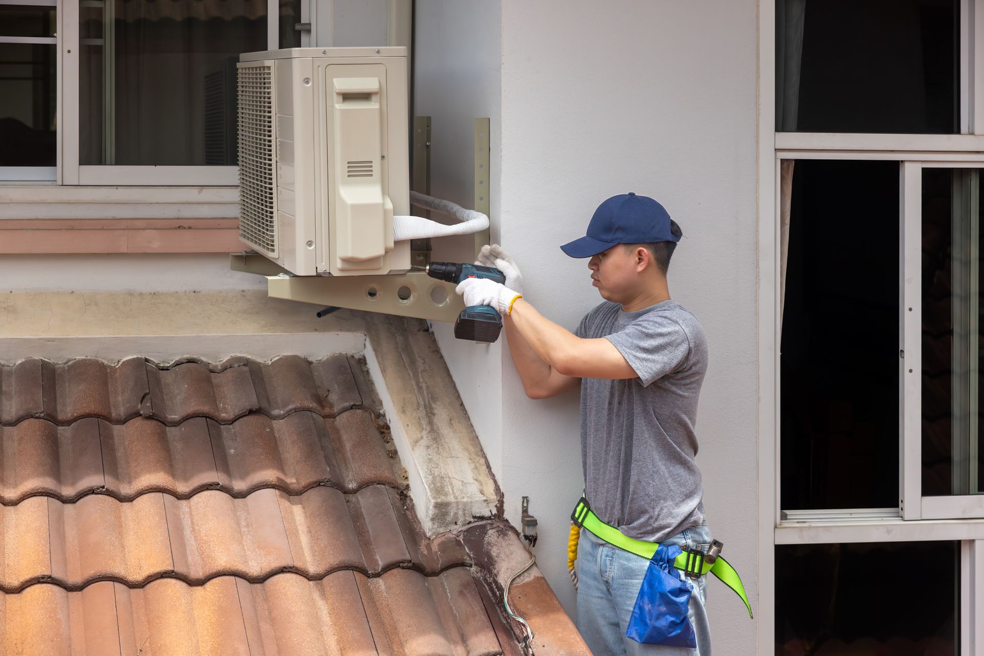 A technician in a blue cap using a drill to install an air conditioning unit on an exterior wall near a tiled roof.