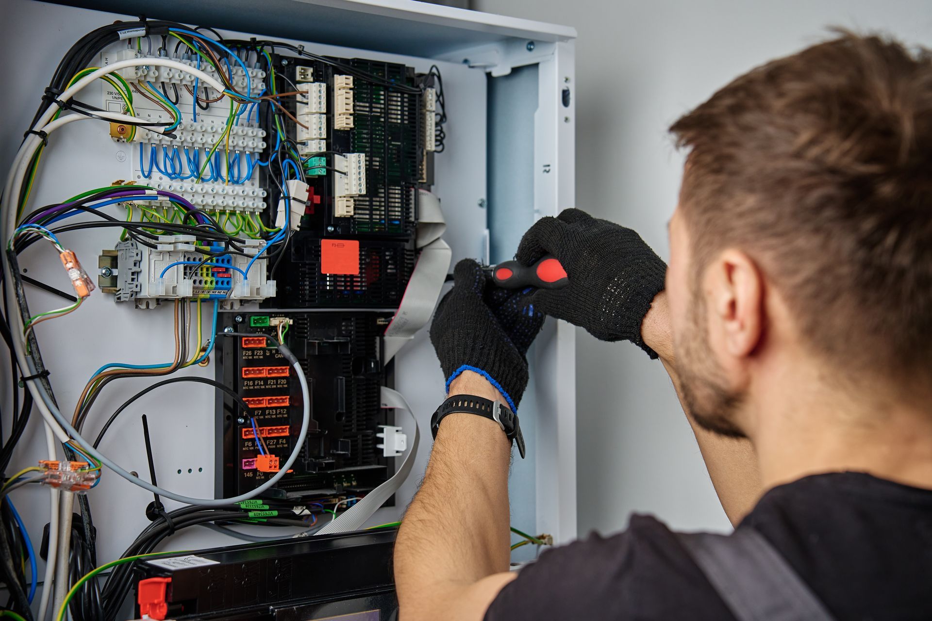 A person wearing black gloves uses a screwdriver to repair a white electrical control panel filled with wires and circuits. A person wearing black gloves uses a screwdriver to repair a white electrical control panel filled with wires and circuits.