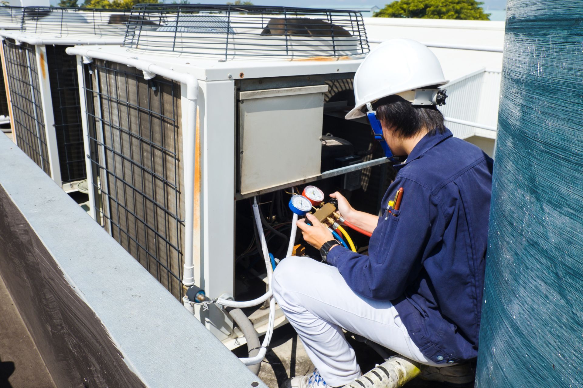 A technician in a hard hat and blue uniform inspects an outdoor air conditioning unit with a manifold gauge set.