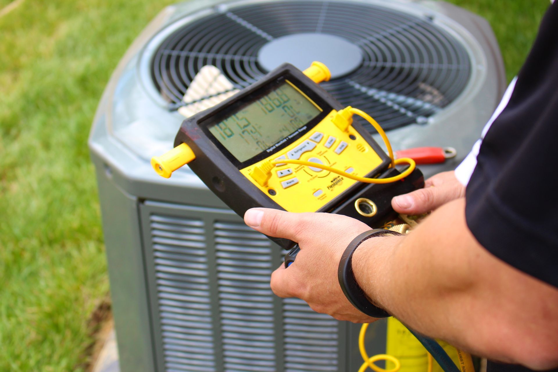 A technician holds a yellow digital HVAC manifold gauge while testing an outdoor air conditioning unit.