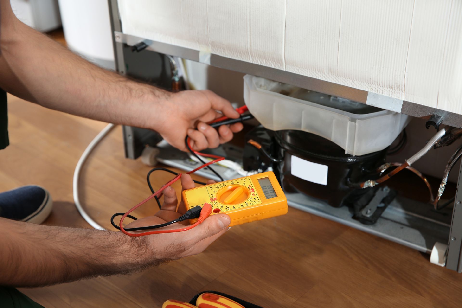 Technician uses a yellow multimeter to test components on the back of a refrigerator near the compressor.