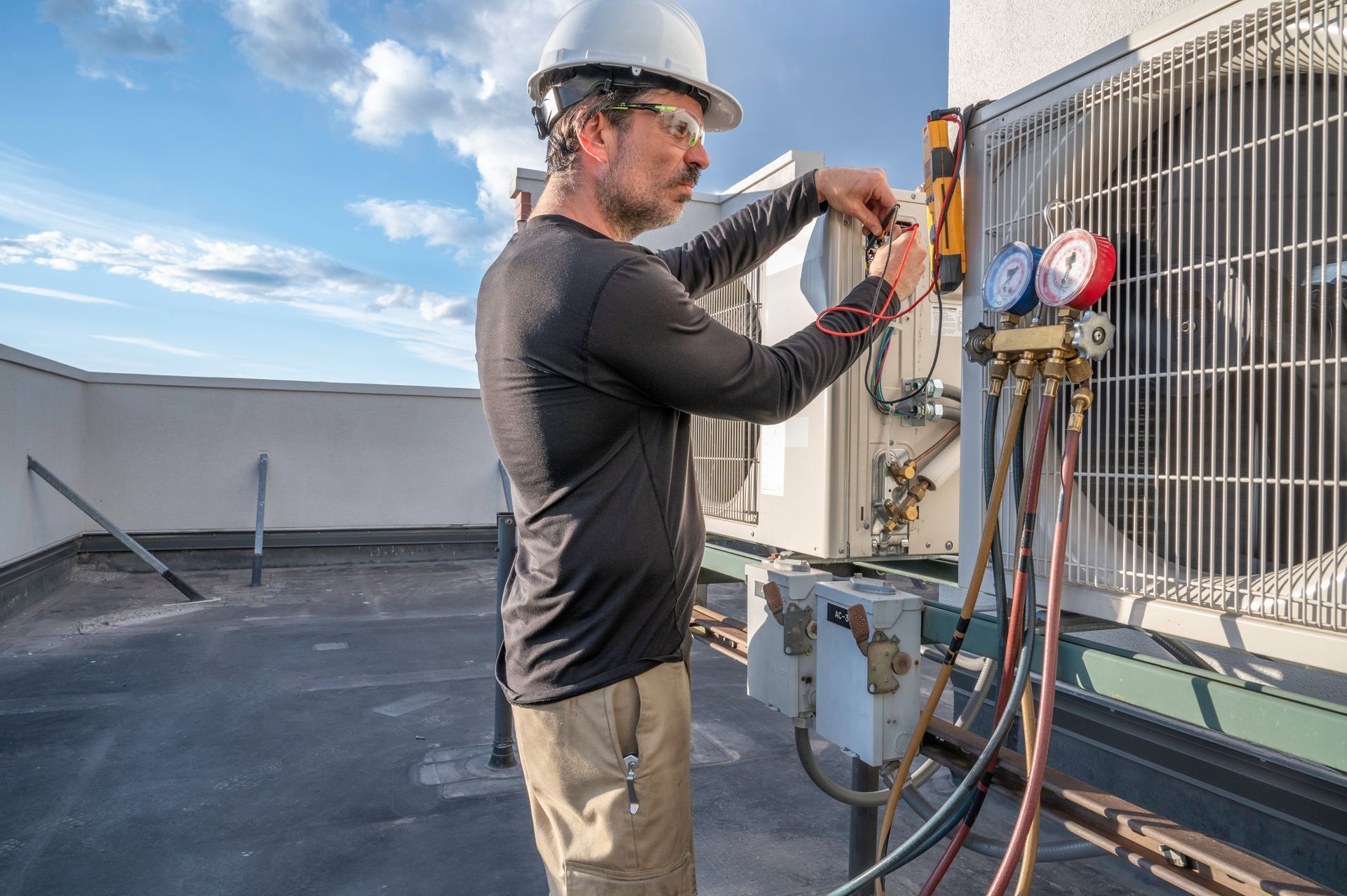 A technician in a white hard hat checks an HVAC unit on a rooftop using a gauge manifold connected to its valves.