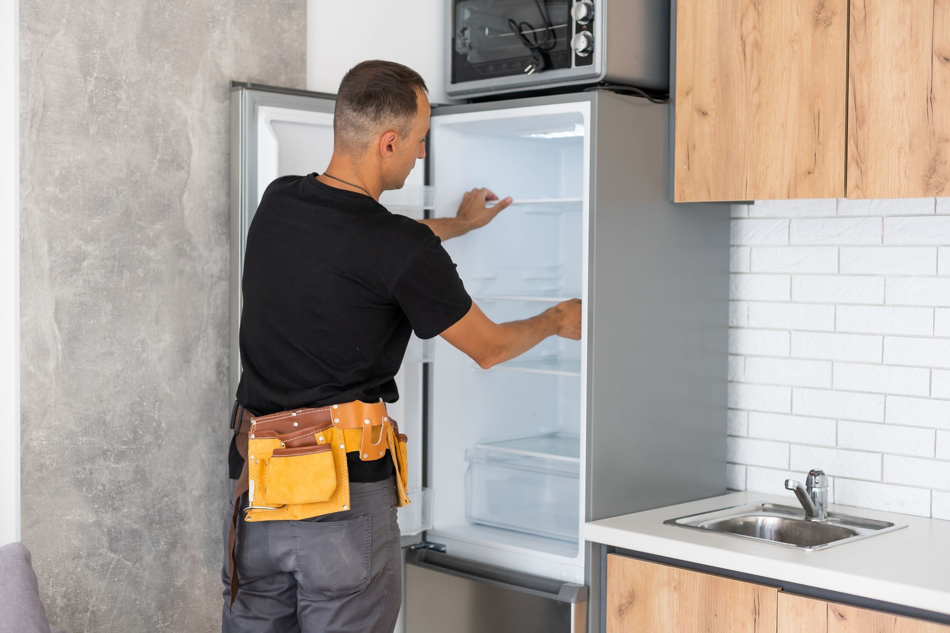 A technician wearing a tool belt inspects the interior of a tall, silver refrigerator in a modern kitchen.