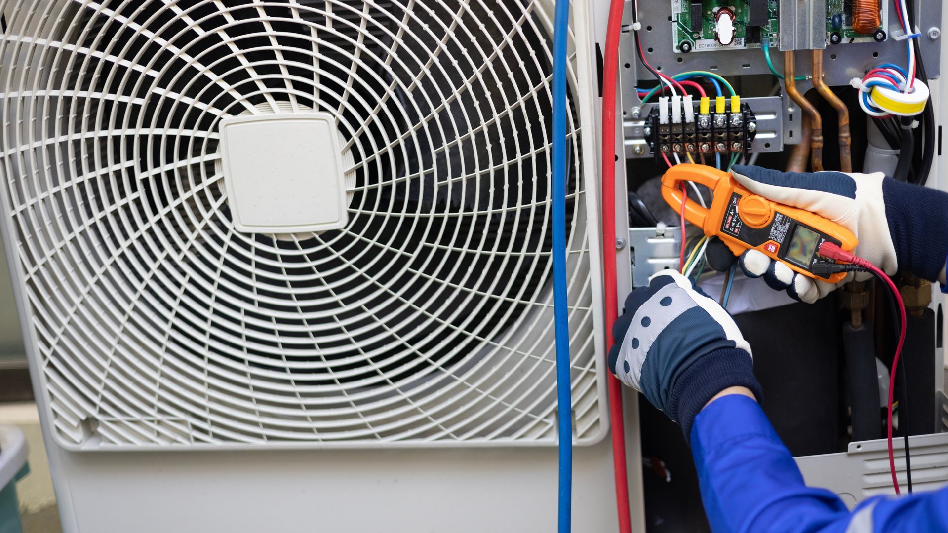 A person wearing work gloves uses a clamp meter to test electrical wiring inside an outdoor air conditioning unit. A person wearing work gloves uses a clamp meter to test electrical wiring inside an outdoor air conditioning unit.