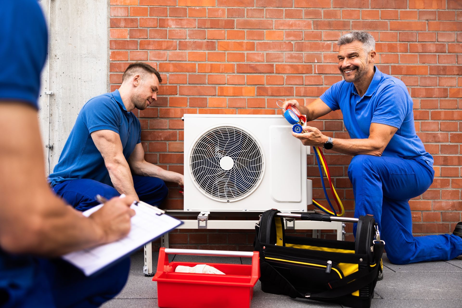 Technicians in blue uniforms inspect an outdoor HVAC unit against a brick wall, using tools and checking a clipboard. Technicians in blue uniforms inspect an outdoor HVAC unit against a brick wall, using tools and checking a clipboard.
