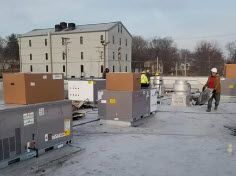 A repair technician in blue overalls kneeling on a kitchen floor while fixing the base of a refrigerator with a tool.