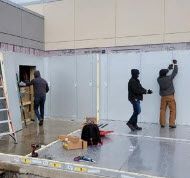 A repair person in blue work overalls inspecting the interior of an open stainless steel refrigerator in a white kitchen.