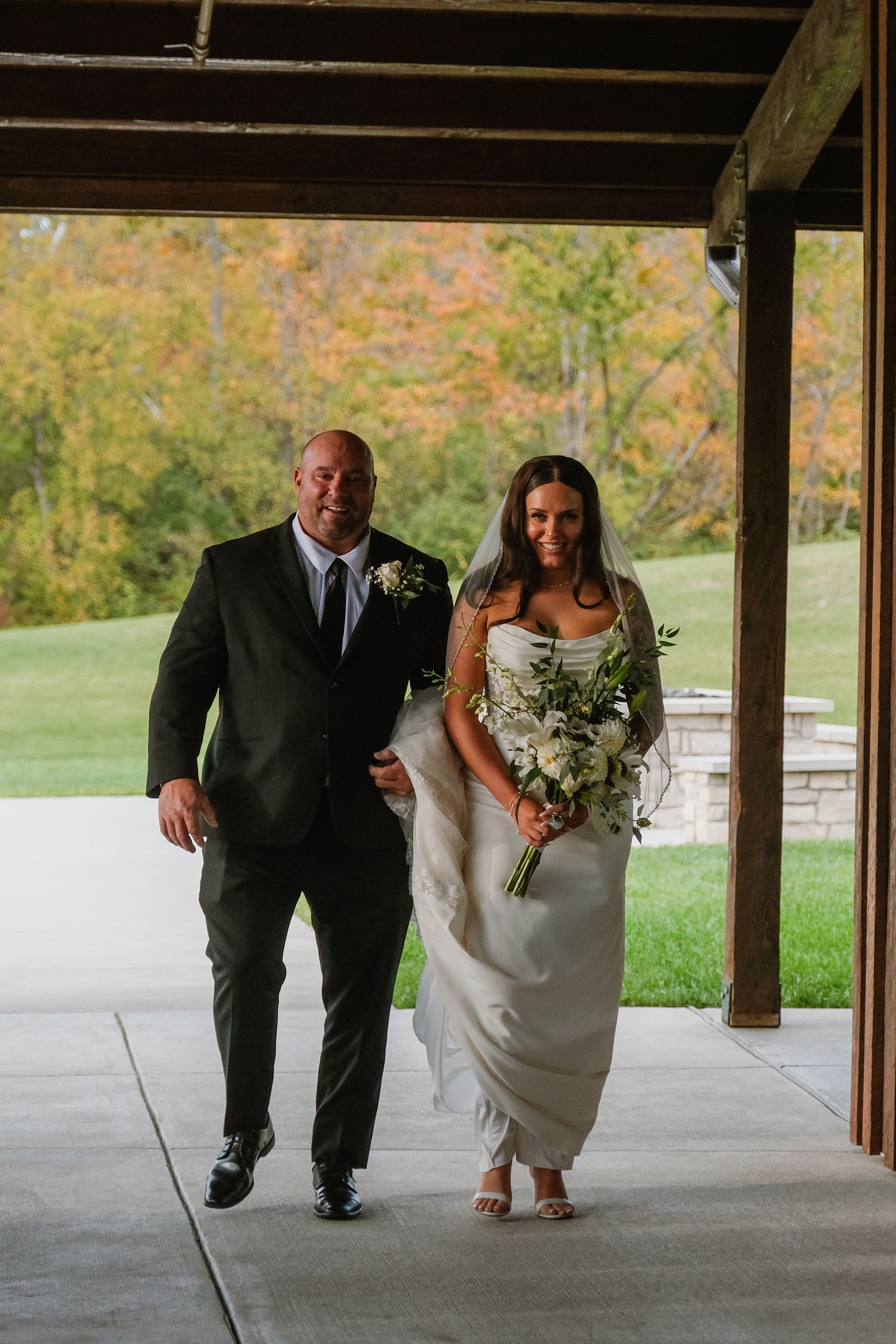 Bride and person escorting her walk down an aisle. Green lawn and fall foliage in the background.