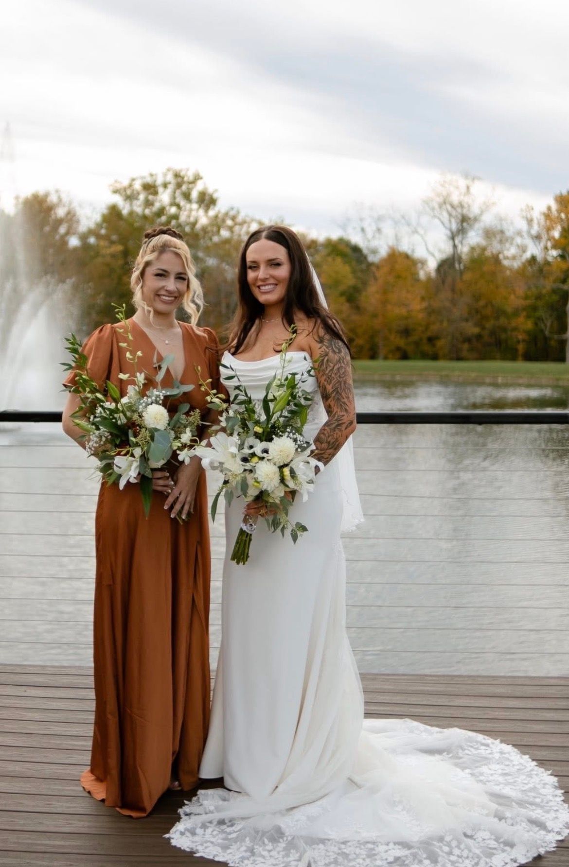 Bride and bridesmaid pose by a lake. Bride in white gown with lace train; bridesmaid in rust dress, both hold bouquets.
