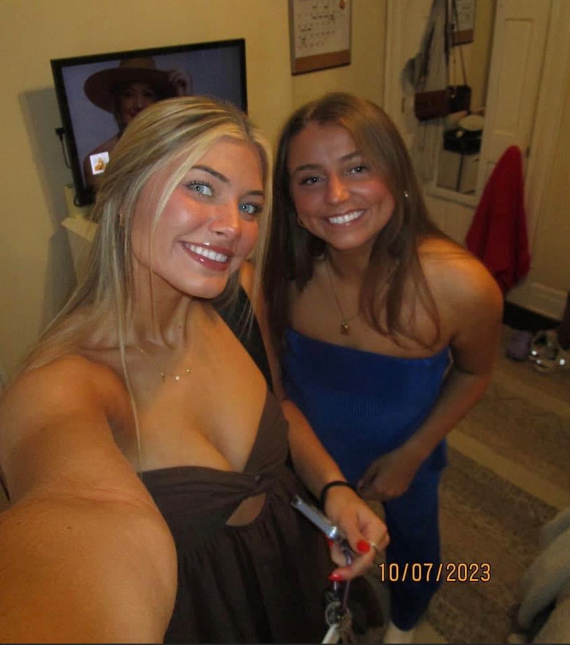Two smiling women take a selfie indoors. One wears a brown dress, the other a blue strapless top. October 7, 2023.