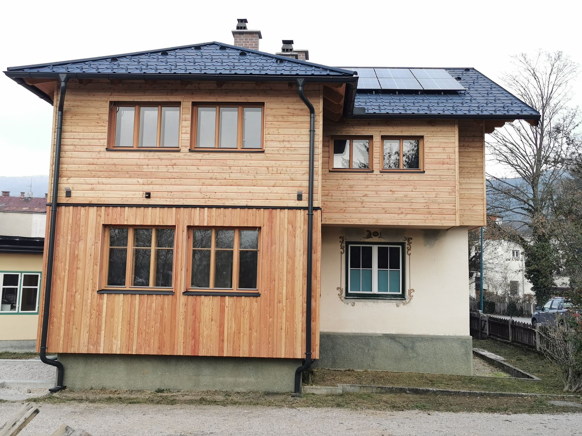 Zweistöckiges Haus mit Holz- und beigefarbener Fassade, dunklem Dach und Solarpaneelen auf dem oberen Dach.