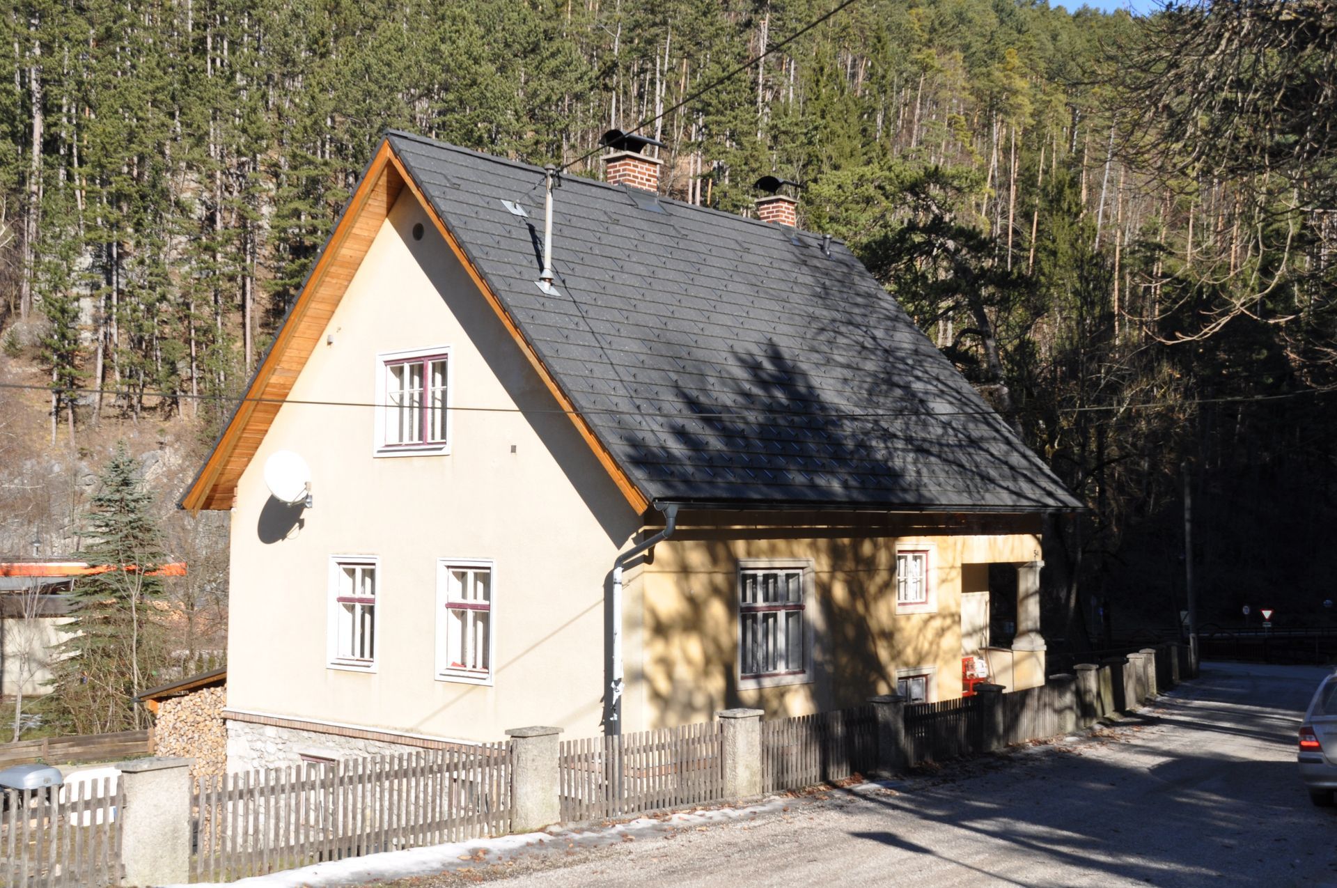 Weißes Haus mit steilem, dunklem Dach neben einer bewaldeten Straße im Sonnenlicht